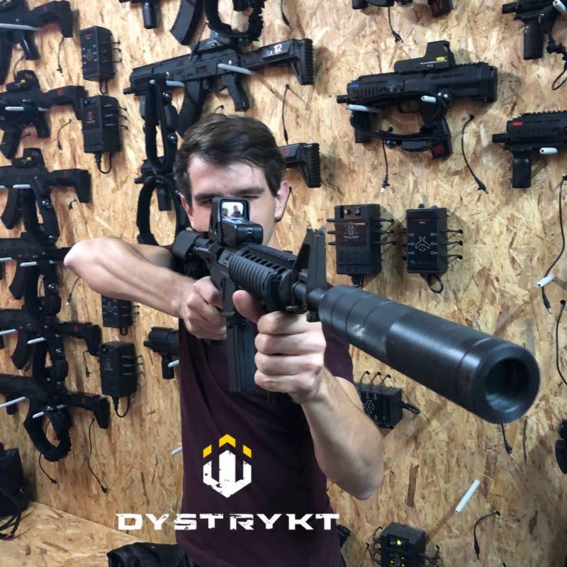 Mężczyzna celuje z karabinu laserowego na tle ściany z rekwizytami do laser tagu, widoczne logo 'DYSTRYKT'.
