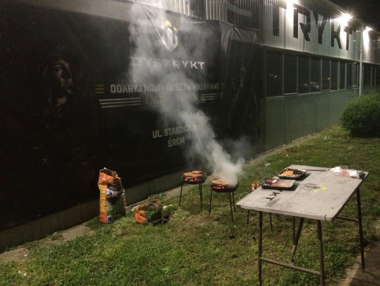 Grillowanie na zewnątrz budynku z banerem reklamowym, unoszący się dym, przygotowane jedzenie na stole.