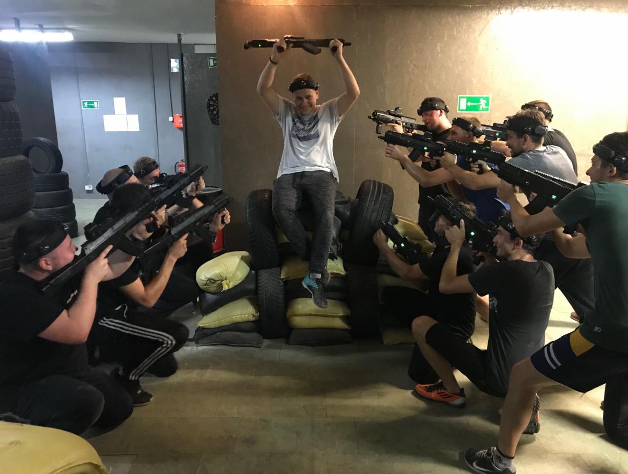 Grupa osób celuje z pistoletów laserowych w pomieszczeniu z oponami i poduszkami; jedna osoba siedzi na stosie opon i trzyma pistolet nad głową.