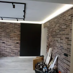 Ściana z dekoracyjnej cegły w odcieniach brązu i szarości z podświetleniem LED przy suficie, czarny panel na ścianie, widoczne gniazdka elektryczne, podłoga z paneli, częściowo widoczne narzędzia...