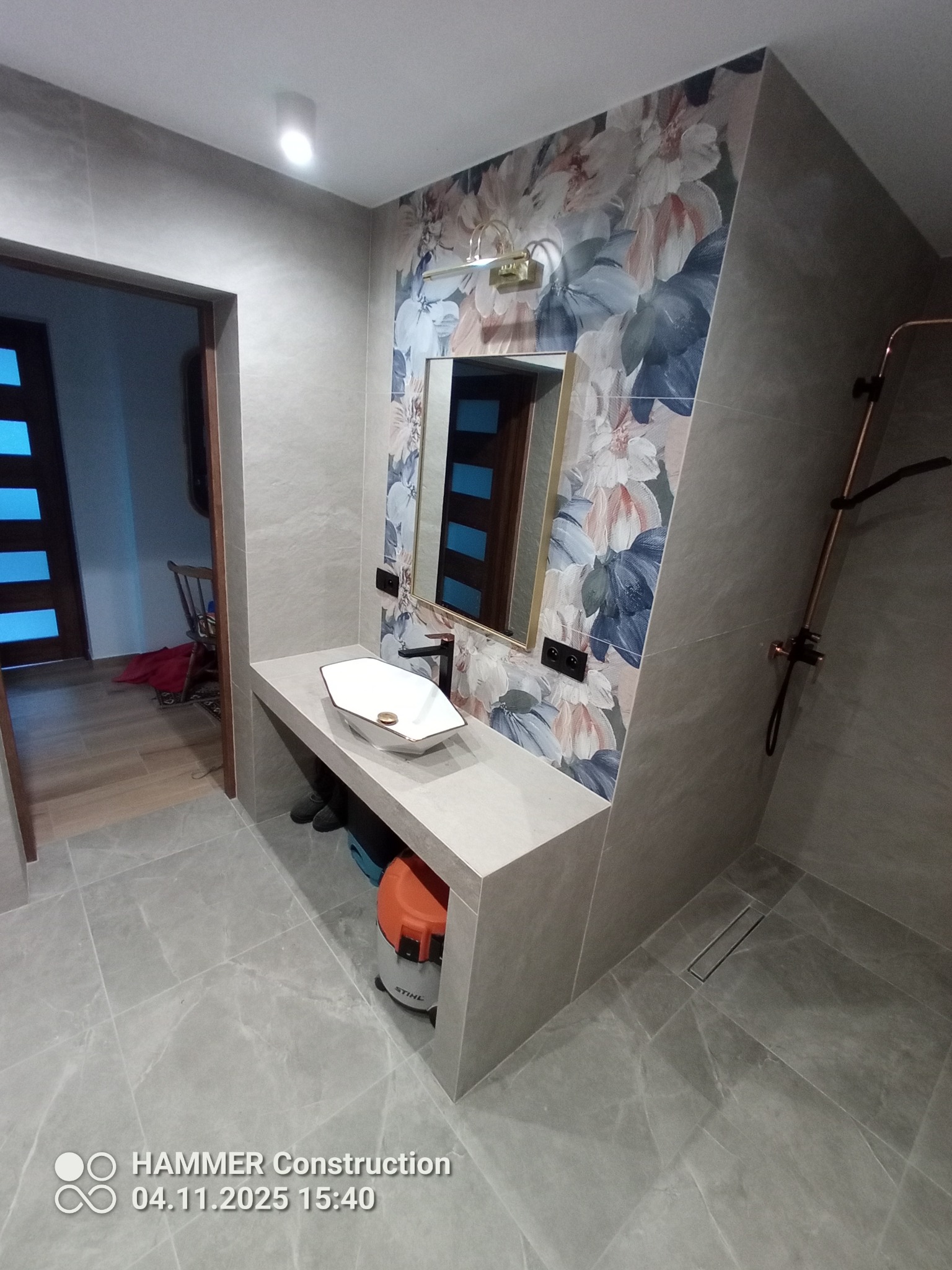 Wykończona łazienka z umywalką nablatową, lustrem w złotej ramie i ścianą w kwiatowe wzory. Szare płytki podłogowe i prysznic bez brodzika z odpływem liniowym.