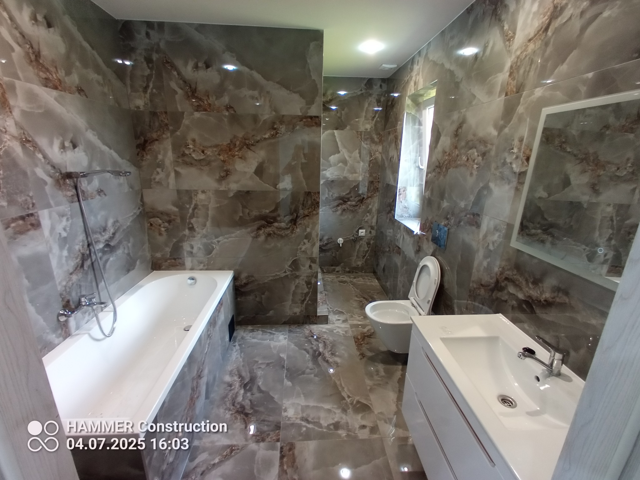 Wykończona łazienka z marmurowymi płytkami na ścianach i podłodze, białą wanną, umywalką z szafką i toaletą. Nowoczesny design z oknem i lustrem.