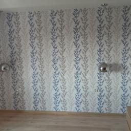 Tegulatoris - Ściana w pokoju wykończona tapetą w pionowe wzory roślinne w odcieniach beżu i niebieskiego, oświetlona przez dwie chromowane lampy wiszące, widoczny fragment drewnianej podłogi i listwy...