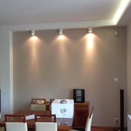 Tegulatoris - Wnętrze pokoju po malowaniu ścian na beżowo, z widocznymi trzema punktowymi lampami sufitowymi, drewnianym stołem z krzesłami i elementami dekoracyjnymi.