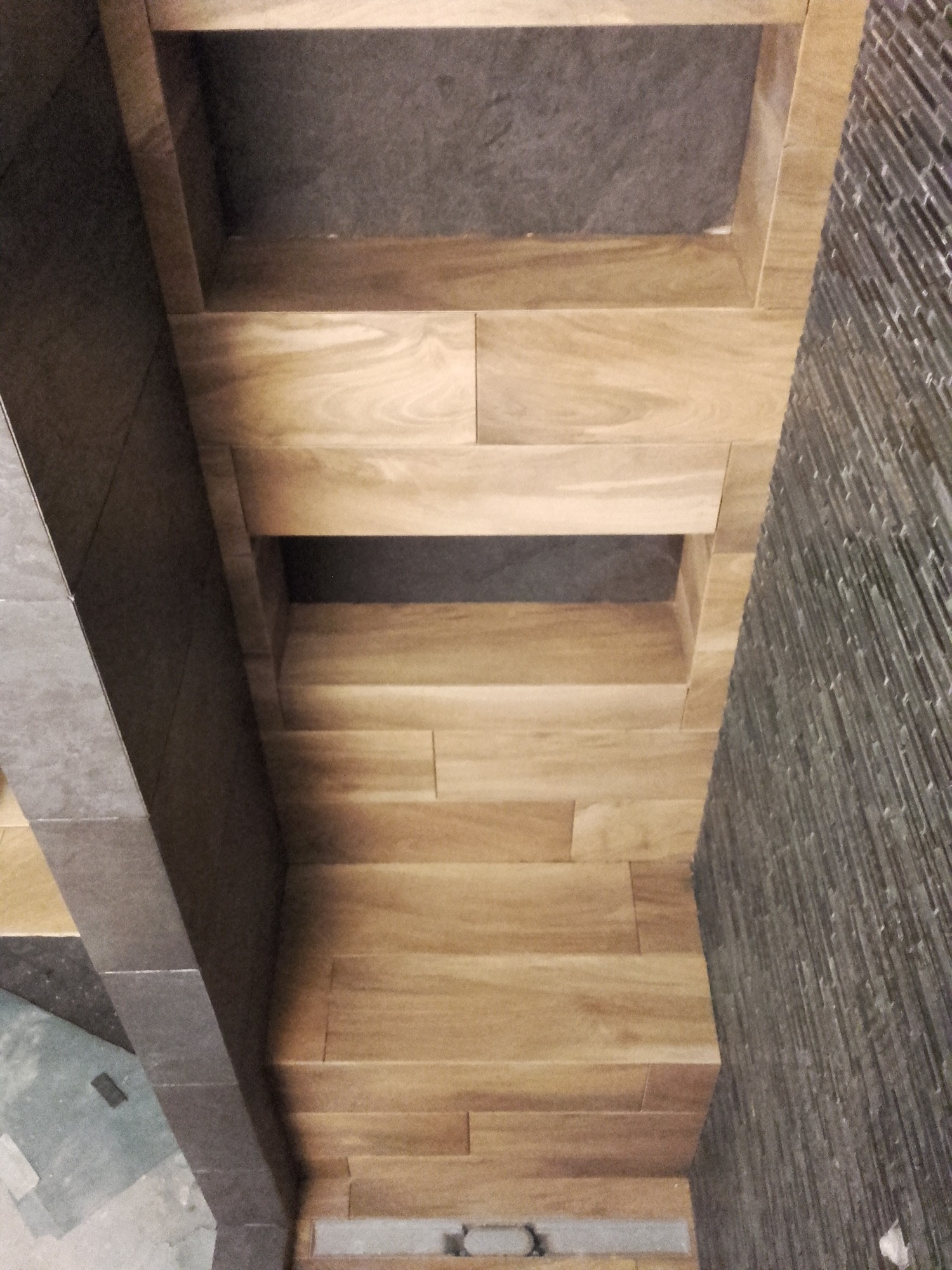 Narożna zabudowa z jasnych, drewnopodobnych płytek z wnękami na ścianie wykończonej ciemną, wąską mozaiką.