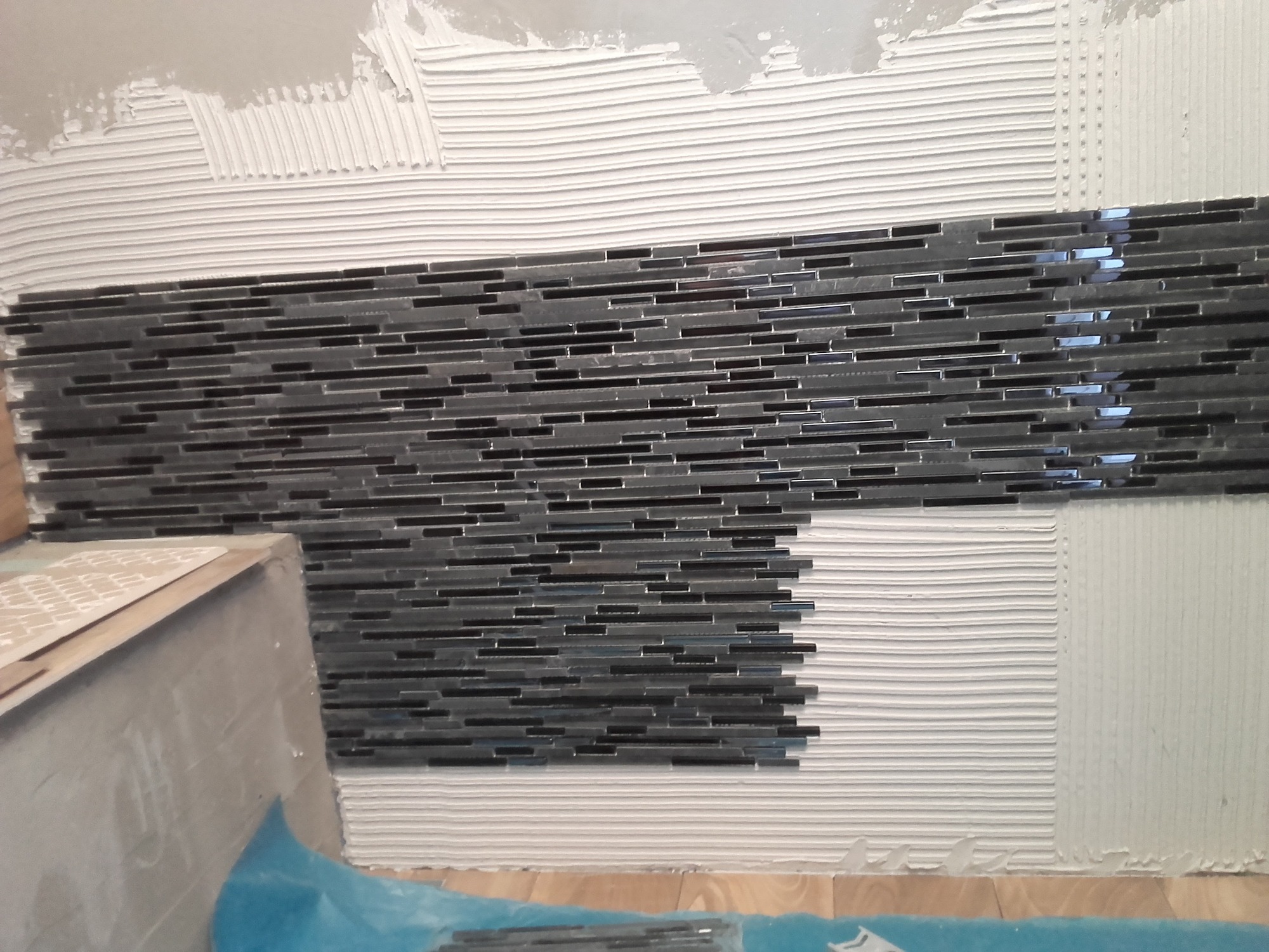 Układanie mozaiki z drobnych, czarnych płytek na ścianie pokrytej świeżym klejem, widoczne niedokończone fragmenty i zabezpieczona folią podłoga.