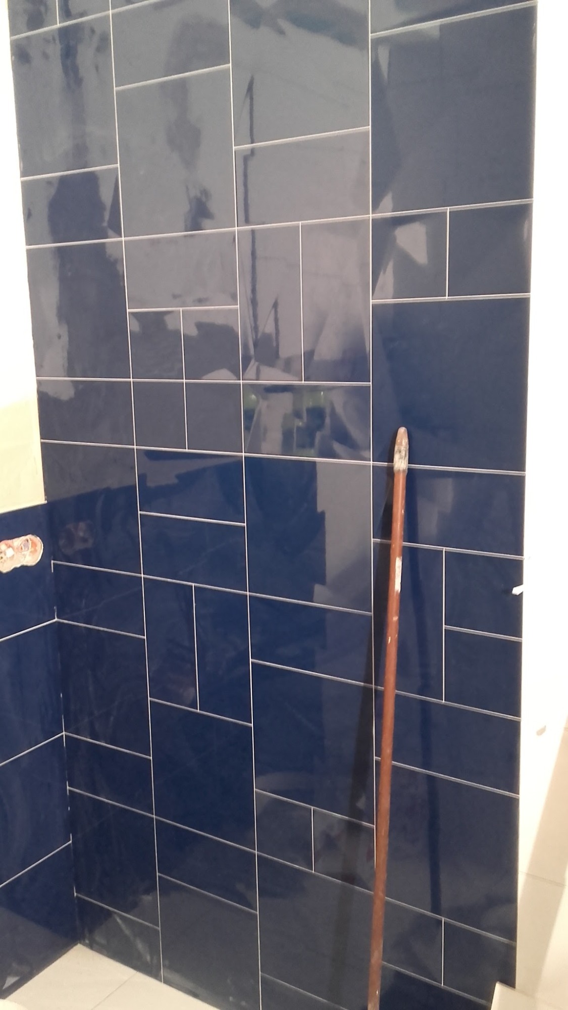 Ściana łazienki wyłożona błyszczącymi, ciemnoniebieskimi płytkami ceramicznymi w układzie poziomym, z widoczną fugą w kolorze białym oraz fragmentem narzędzia glazurniczego w rogu.