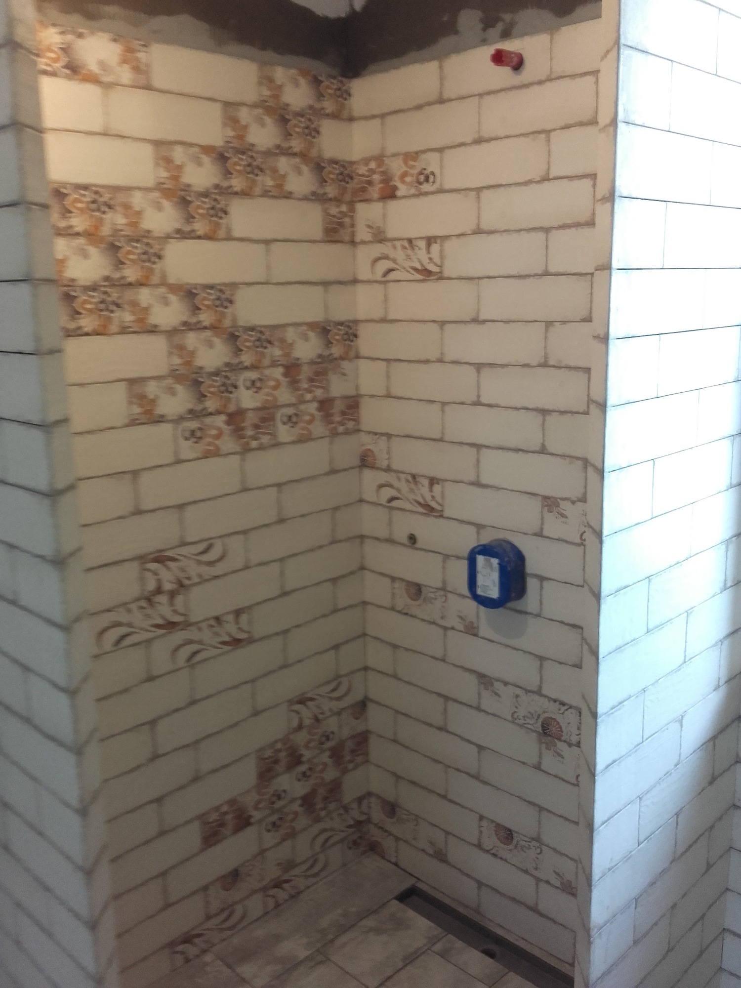 Świeżo ułożone, beżowo-białe kafelki w kształcie cegiełek, z dekoracyjnymi wstawkami w odcieniach brązu i pomarańczy, w narożnej kabinie prysznicowej z odpływem liniowym w podłodze i niebieskim...