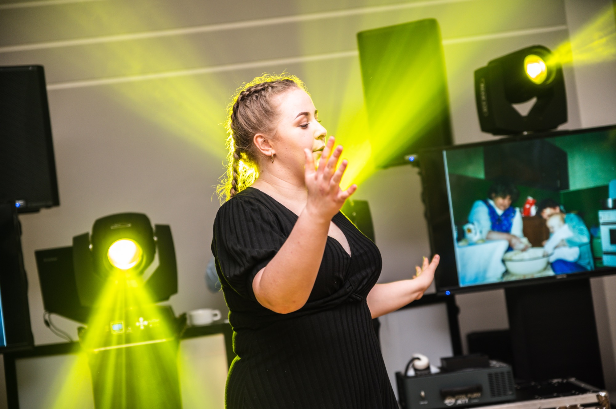 Kobieta w czarnej sukience gestykuluje na tle sceny z oświetleniem eventowym i ekranem wyświetlającym zdjęcie rodzinne.