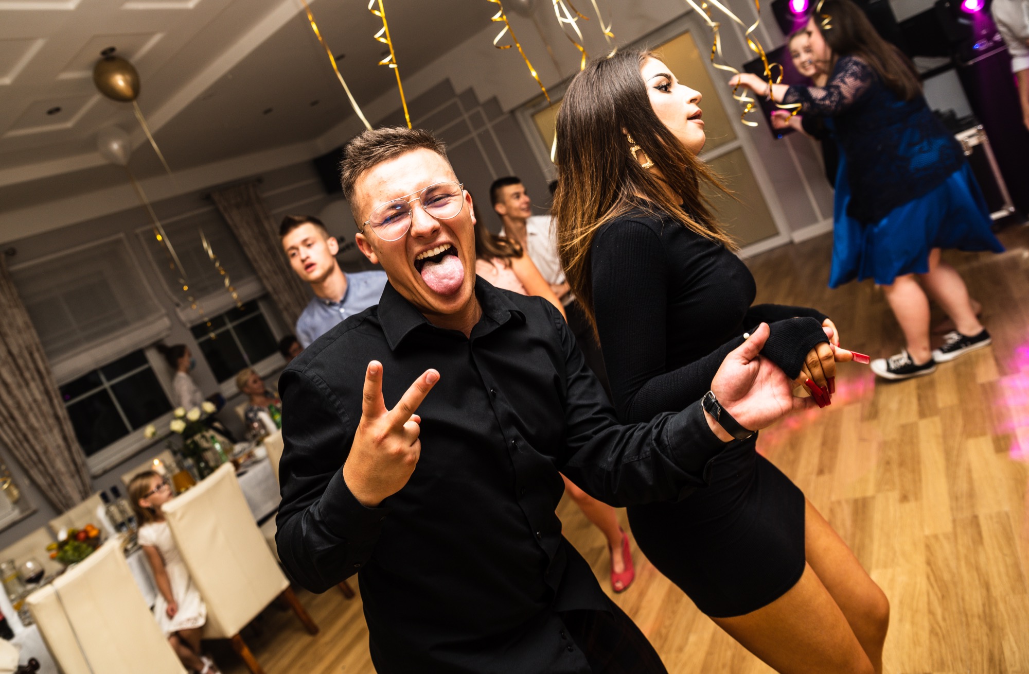Rozbawiony mężczyzna w okularach pokazuje język i gest zwycięstwa na parkiecie podczas eventu, obok tańcząca kobieta w czarnej sukience, w tle bawiący się goście i dekoracje.