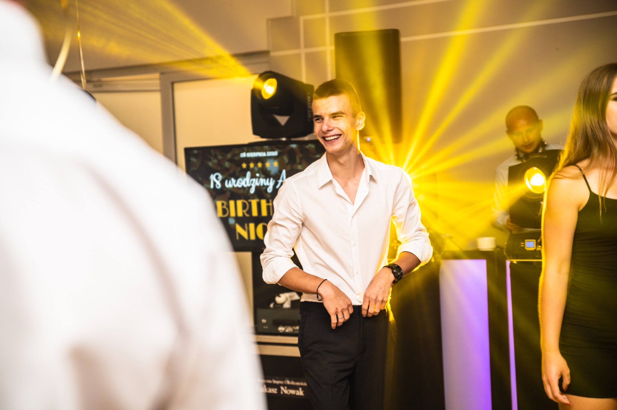Uśmiechnięty młody mężczyzna w białej koszuli na imprezie urodzinowej, w tle DJ i oświetlenie sceniczne.