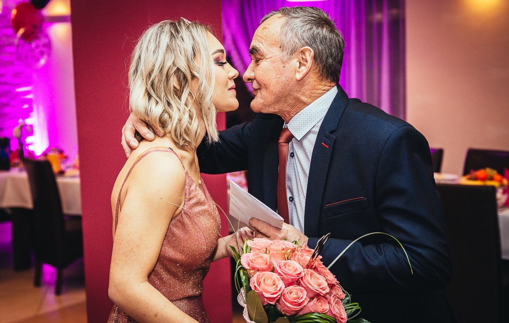 Młoda kobieta w błyszczącej sukience i starszy mężczyzna w garniturze, trzymający bukiet róż, w czułym uścisku na tle sali bankietowej z dekoracjami.
