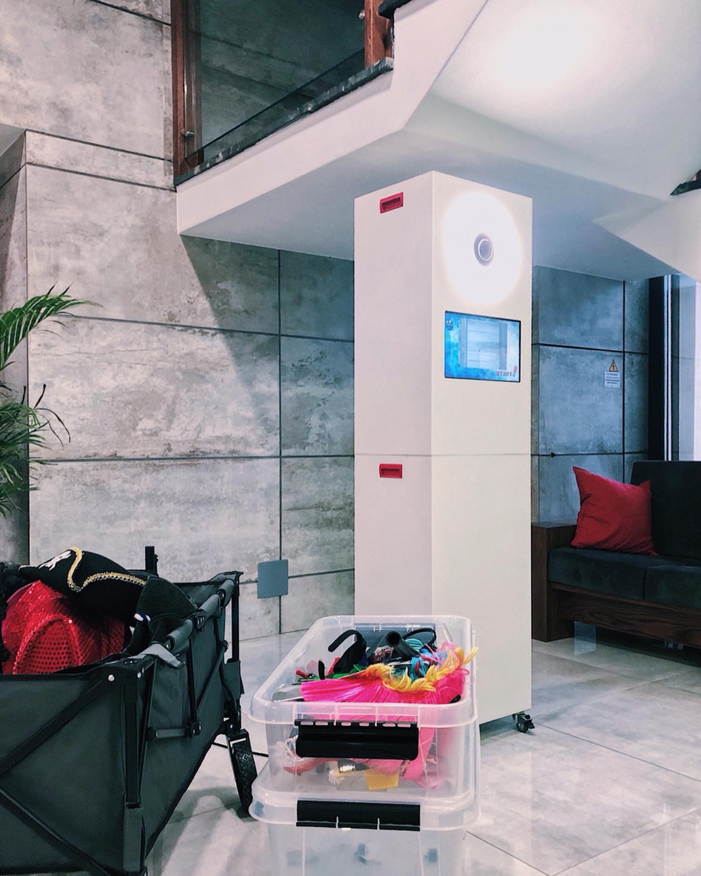 Stanowisko do robienia zdjęć z ekranem dotykowym i rekwizytami w wózku i plastikowym pojemniku, wnętrze z szarą ścianą i sofą z czerwoną poduszką.