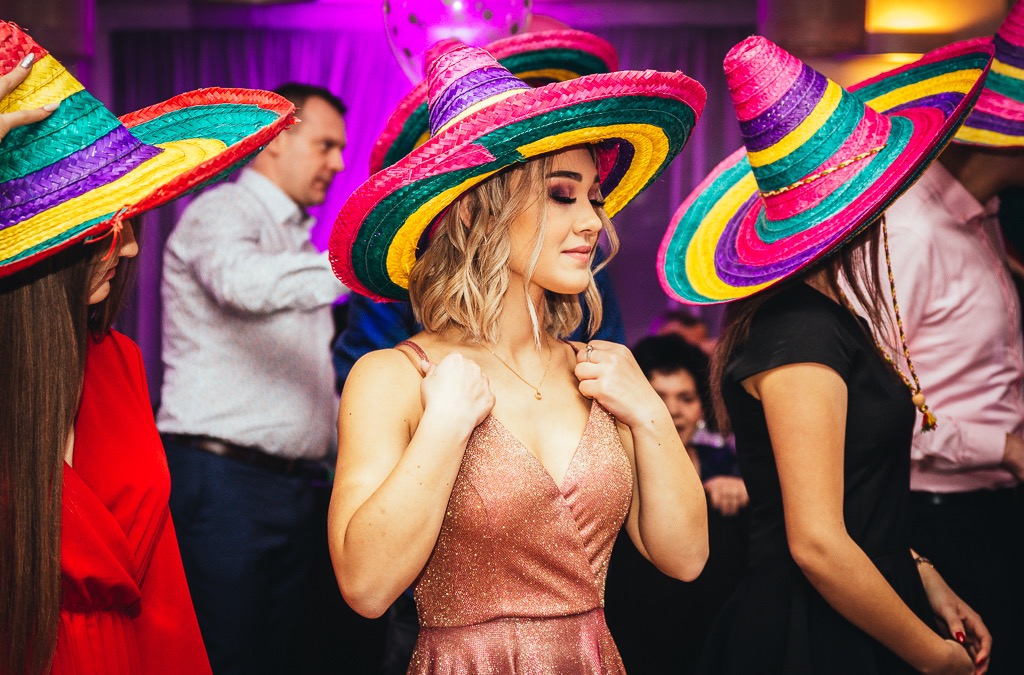 Uczestnicy imprezy w kolorowych sombrerach, kobieta w błyszczącej sukience i złotym naszyjniku, uśmiechnięta.