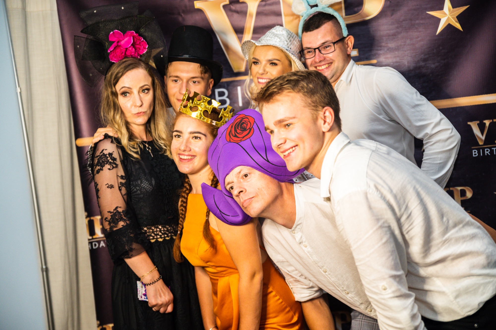 Grupa osób pozuje do zdjęcia na tle dekoracji urodzinowej VIP, ubrani w zabawne kapelusze i przebrania.