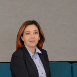 Portret uśmiechniętej kobiety z rudymi włosami, w czarnej marynarce i błękitnej koszuli w kropki, siedzącej na turkusowej sofie na tle szarej ściany.