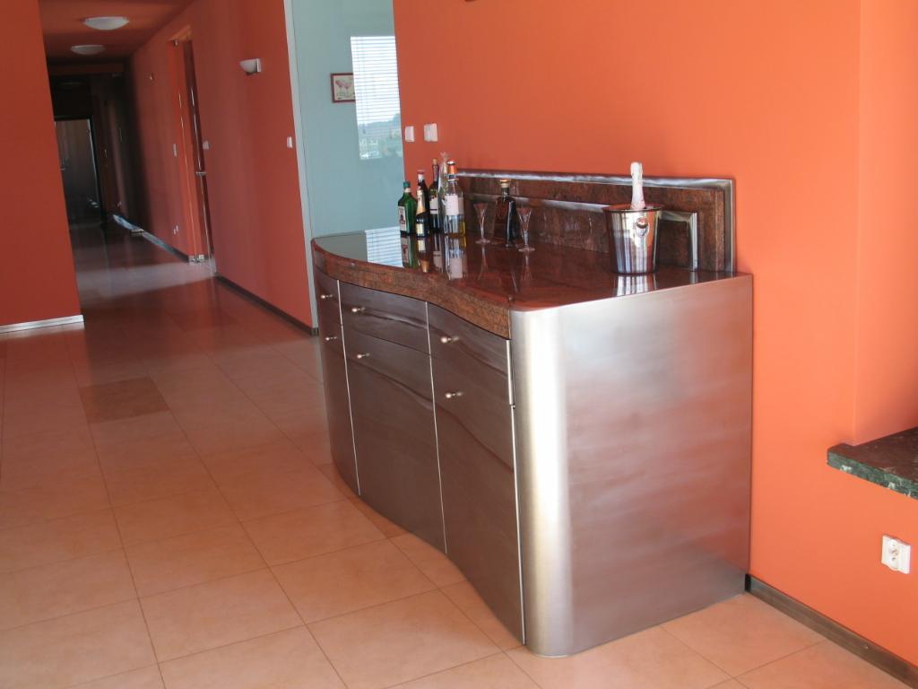 Nowoczesna komoda z blatem z ciemnego kamienia i frontami z nierdzewnej stali, z butelkami alkoholu i wiaderkiem na szampana na blacie, na tle pomarańczowej ściany i długiego korytarza.