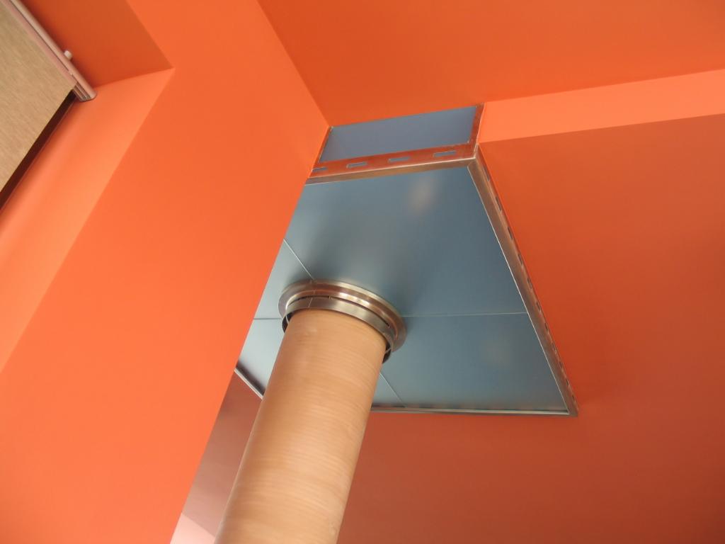 Fragment instalacji wentylacyjnej z rurą o teksturowanej powierzchni i metalową obudową, na tle pomarańczowego sufitu i ścian.