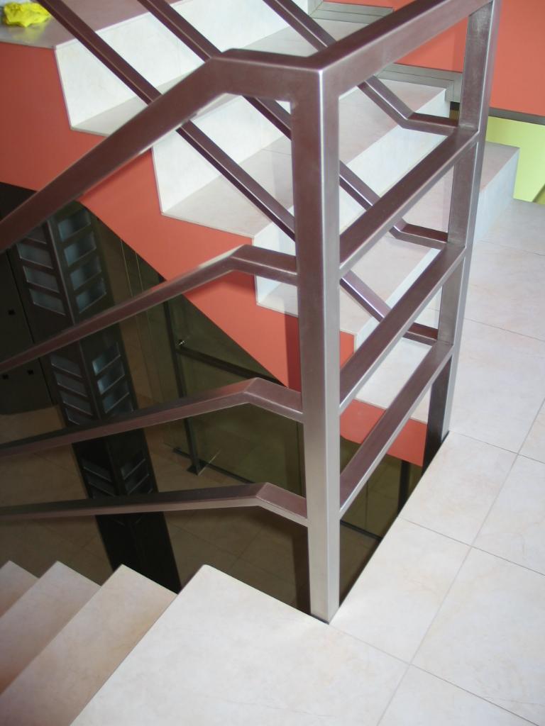 Klatka schodowa z kremowymi stopniami i geometryczną metalową barierką w odcieniach szarości, na tle pomarańczowej ściany i kafelkowej podłogi.