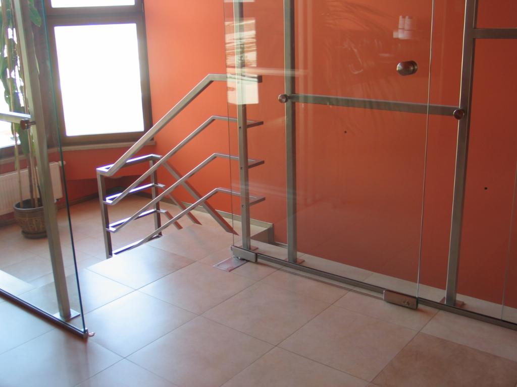 Nowoczesne schody z metalową konstrukcją i szklaną ścianą działową, widoczne z boku na tle pomarańczowej ściany.