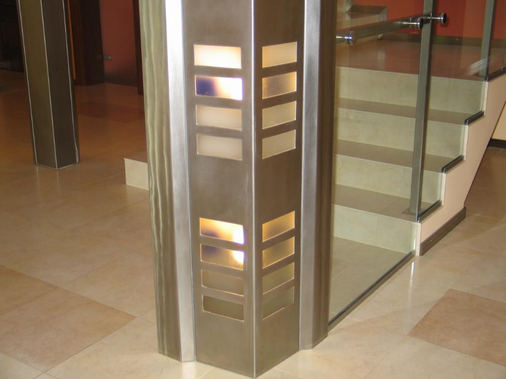 Szczegółowe ujęcie nowoczesnych schodów z szklaną balustradą i metalową kolumną z podświetlanymi, prostokątnymi otworami na beżowej podłodze.