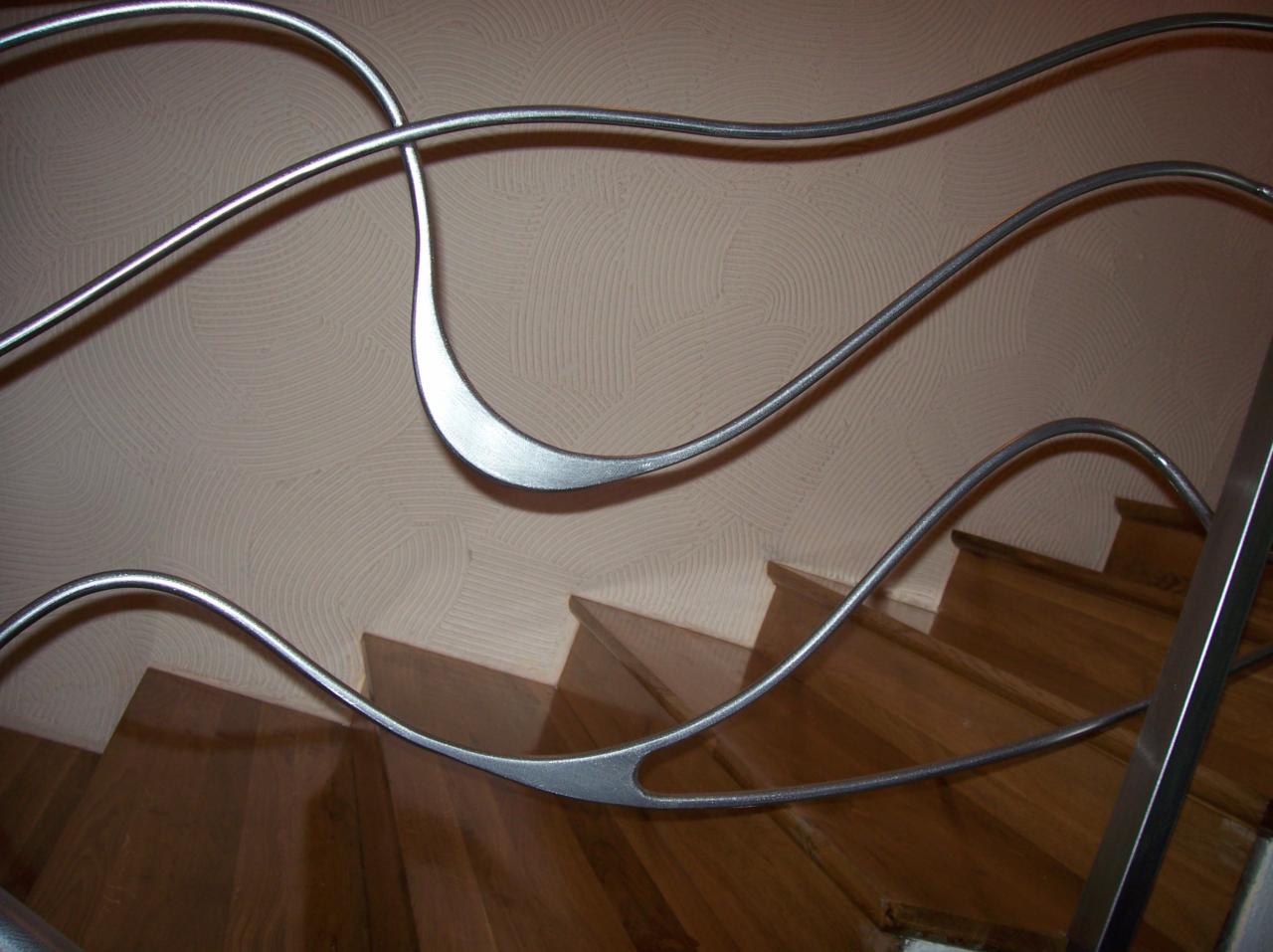 Nietypowa, falująca poręcz ze stali nierdzewnej zamontowana na drewnianych schodach, widok od dołu, beżowa ściana z dekoracyjną fakturą w tle.