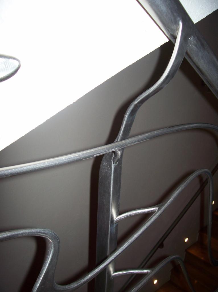 Szczegółowe ujęcie giętej, metalowej konstrukcji zabezpieczającej schody, z widocznymi spawami i oświetleniem punktowym na stopniach.