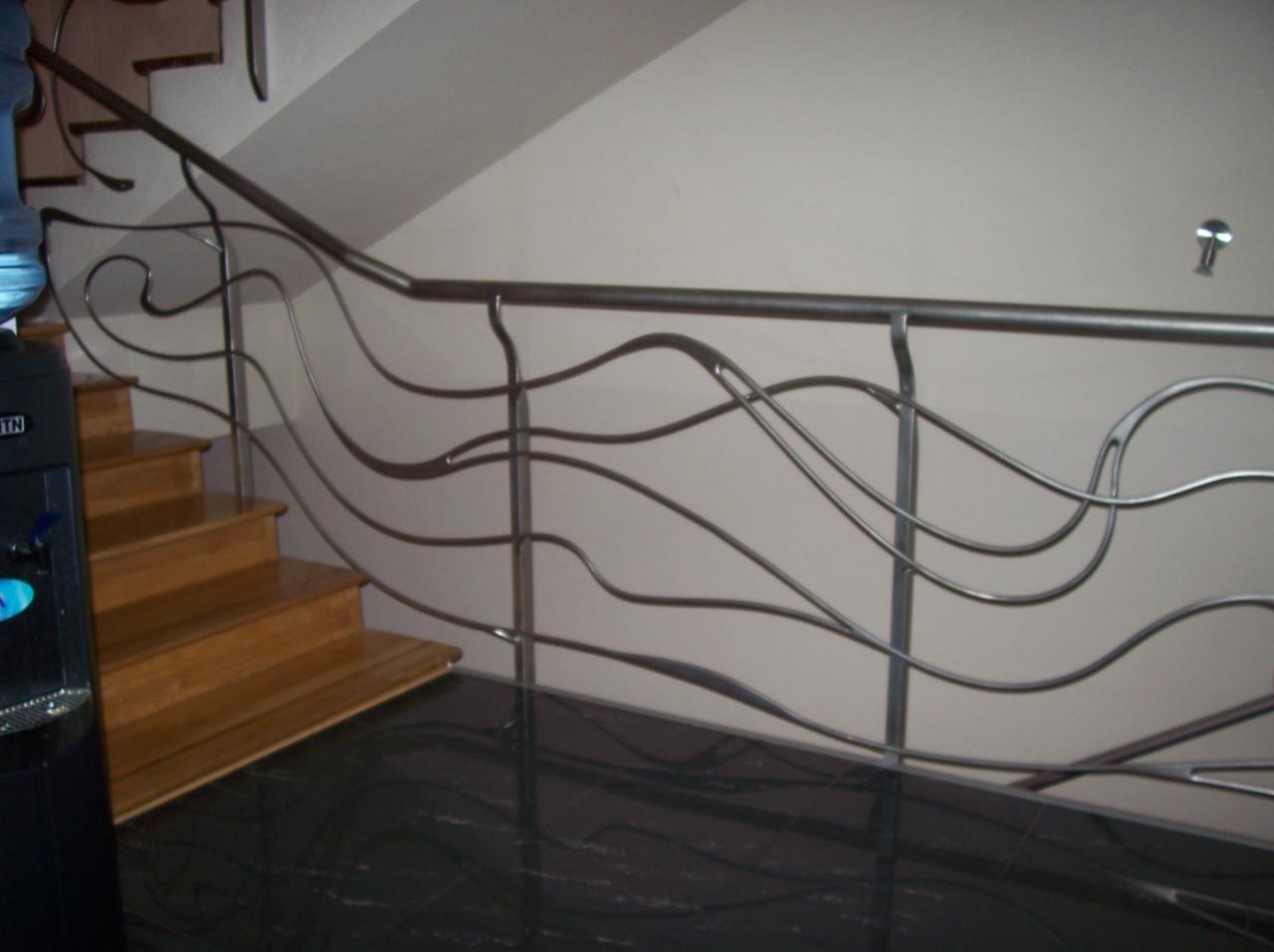 Nowoczesna balustrada ze stali z motywem falistym, zamontowana przy drewnianych schodach, widok z dołu na błyszczącą, ciemną podłogę.