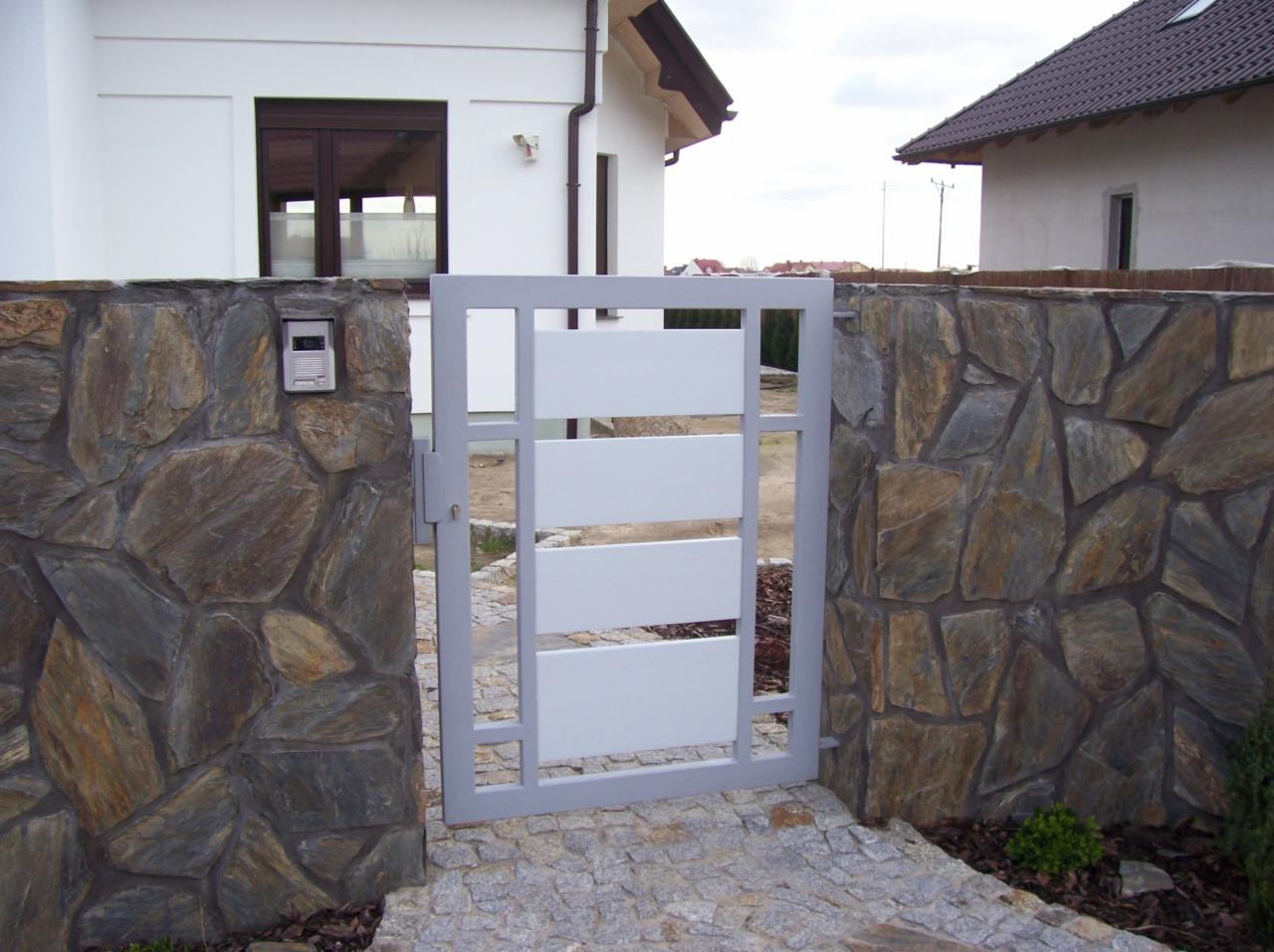 Nowoczesna, biała furtka z poziomymi panelami, osadzona w kamiennym murze z nieregularnych, brązowo-szarych kamieni, prowadząca na ścieżkę z kostki brukowej.