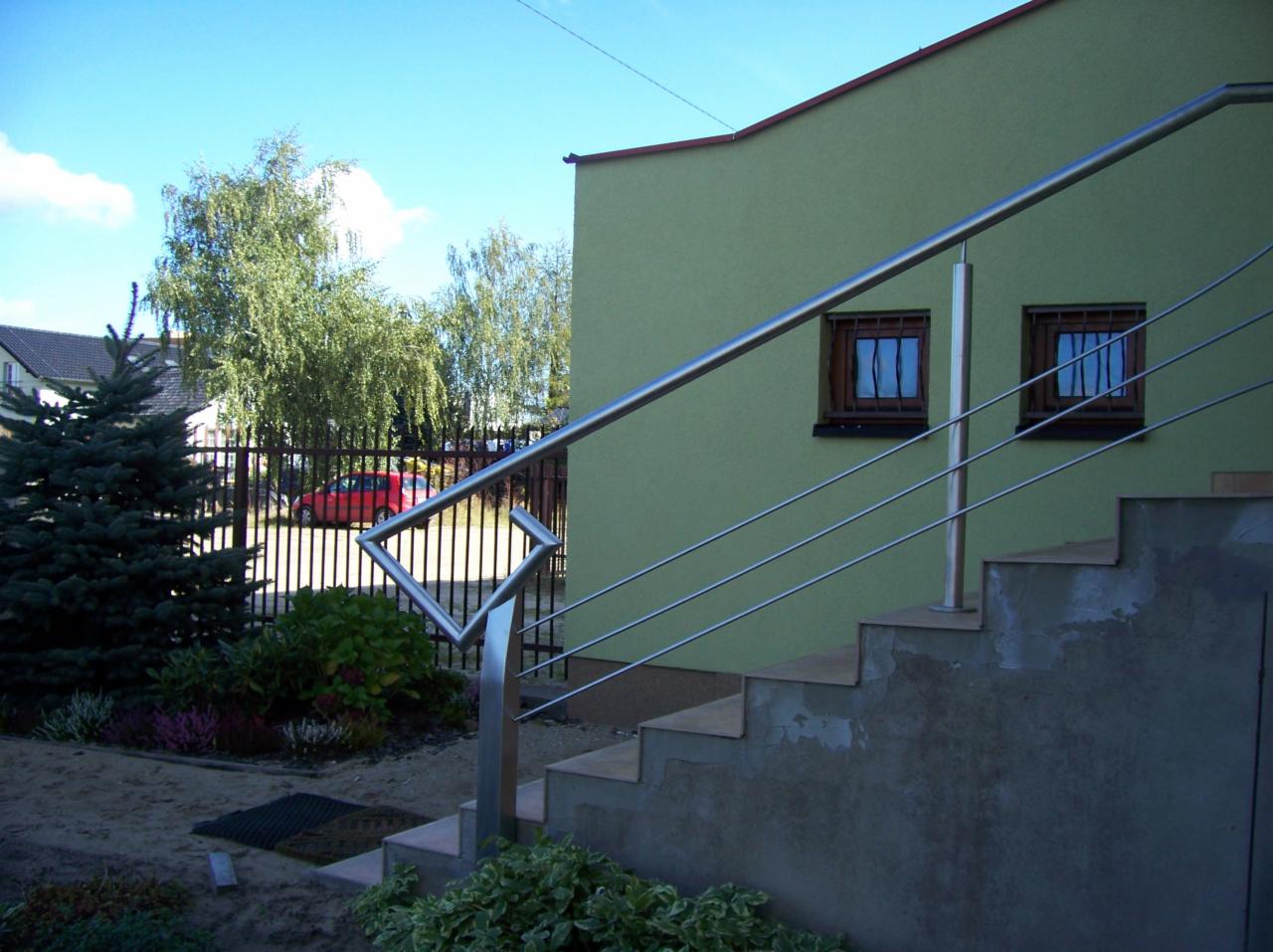 Zewnętrzne schody z szarą betonową podstawą i ze stalową, minimalistyczną barierką z cienkich prętów, na tle zielonej ściany budynku z dwoma oknami z kratami.