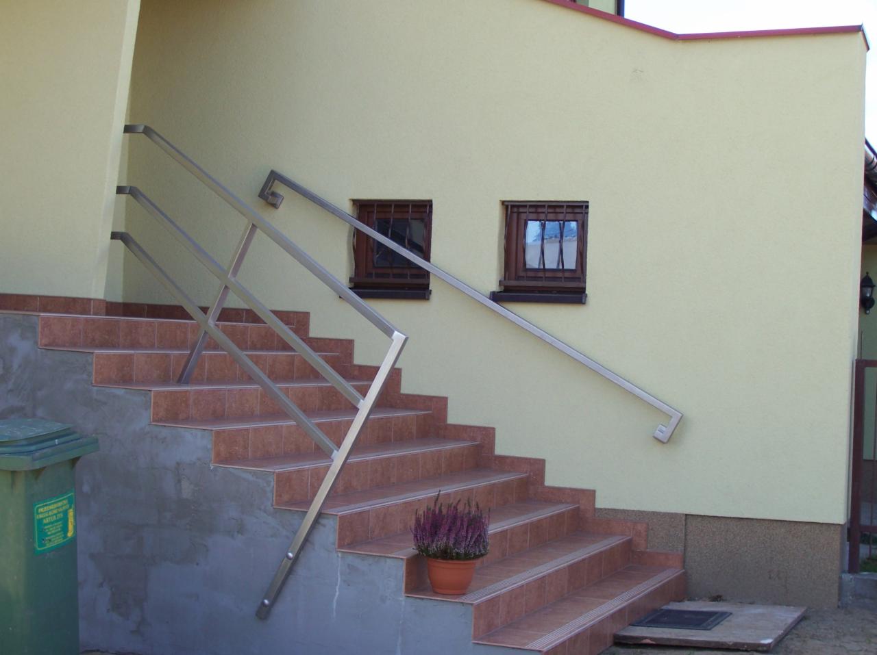 Zewnętrzne schody wyłożone brązowymi płytkami z metalową poręczą, doniczka z wrzosem na jednym ze stopni, obok śmietnik.