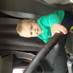 Mały chłopiec w zielonej bluzie bawi się za kierownicą czarnego samochodu ciężarowego, siedząc na szarym siedzeniu. W tle zasłony w kabinie.