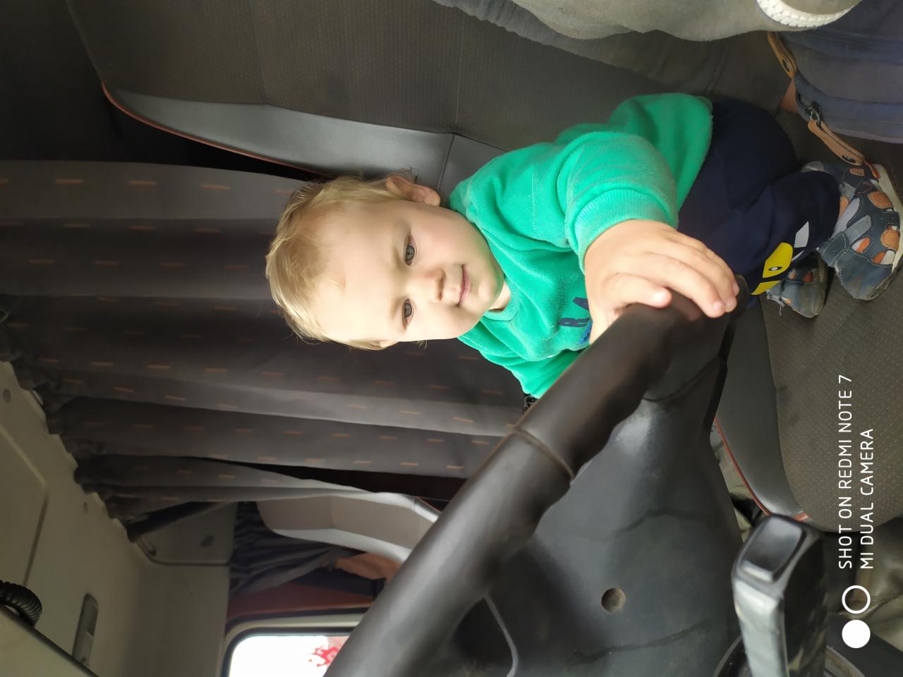 Mały chłopiec w zielonej bluzie bawi się za kierownicą czarnego samochodu ciężarowego, siedząc na szarym siedzeniu. W tle zasłony w kabinie.