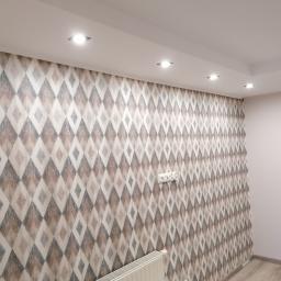 Projekt Venar - Ściana pokryta tapetą w romby w odcieniach beżu, szarości i bieli, z widocznymi gniazdkami elektrycznymi i grzejnikiem pod spodem, oświetlona wpuszczanymi lampami sufitowymi.