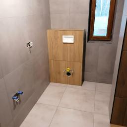 Projekt Venar - Montaż podtynkowego stelaża WC z drewnianą obudową w łazience wyłożonej szarymi płytkami, widoczne wystające rury instalacji wodnej i elektrycznej.