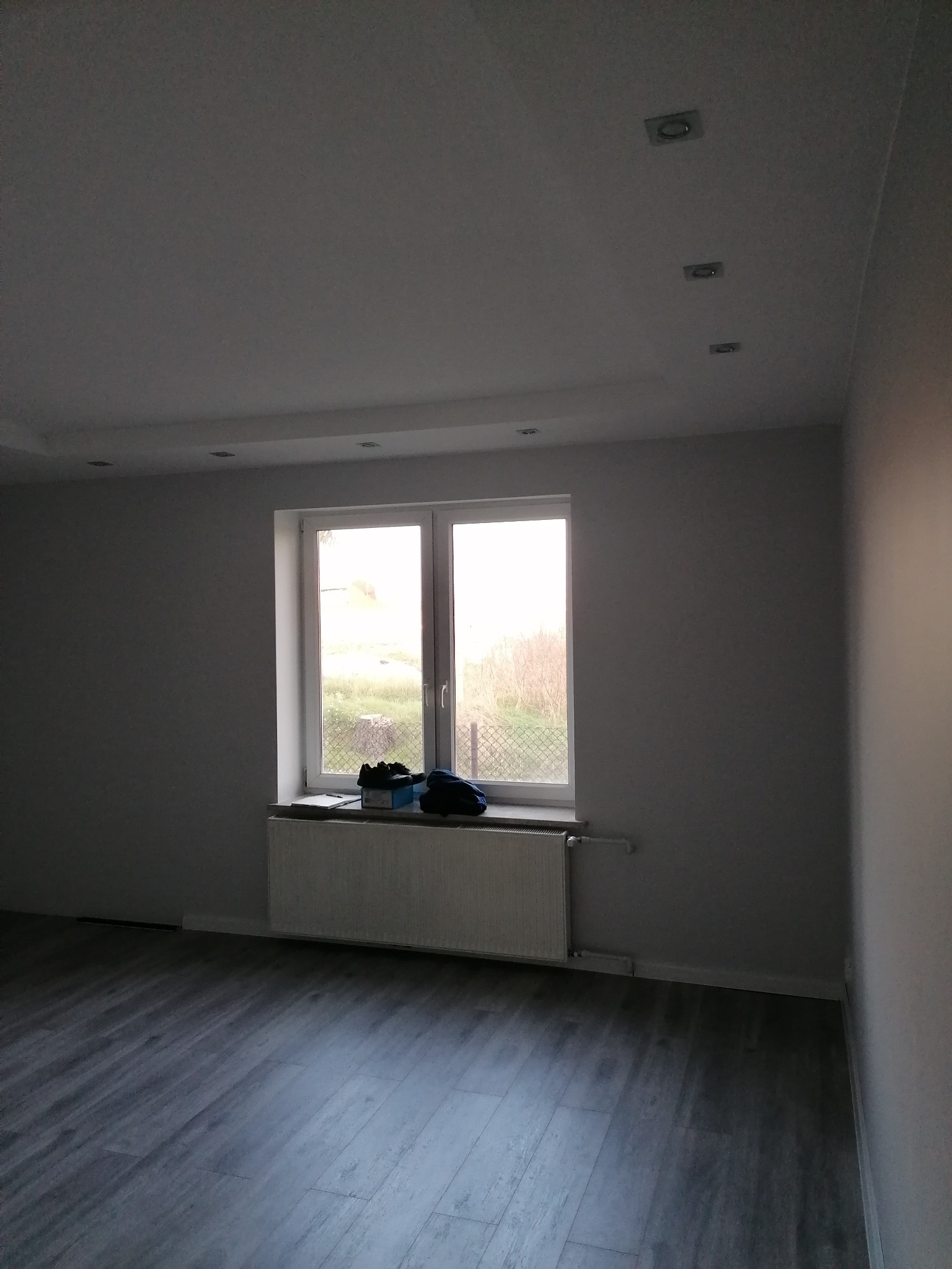 Pomalowane na szaro wnętrze pokoju z panelami podłogowymi, białym oknem i grzejnikiem. Na parapecie leżą buty i pudełko.