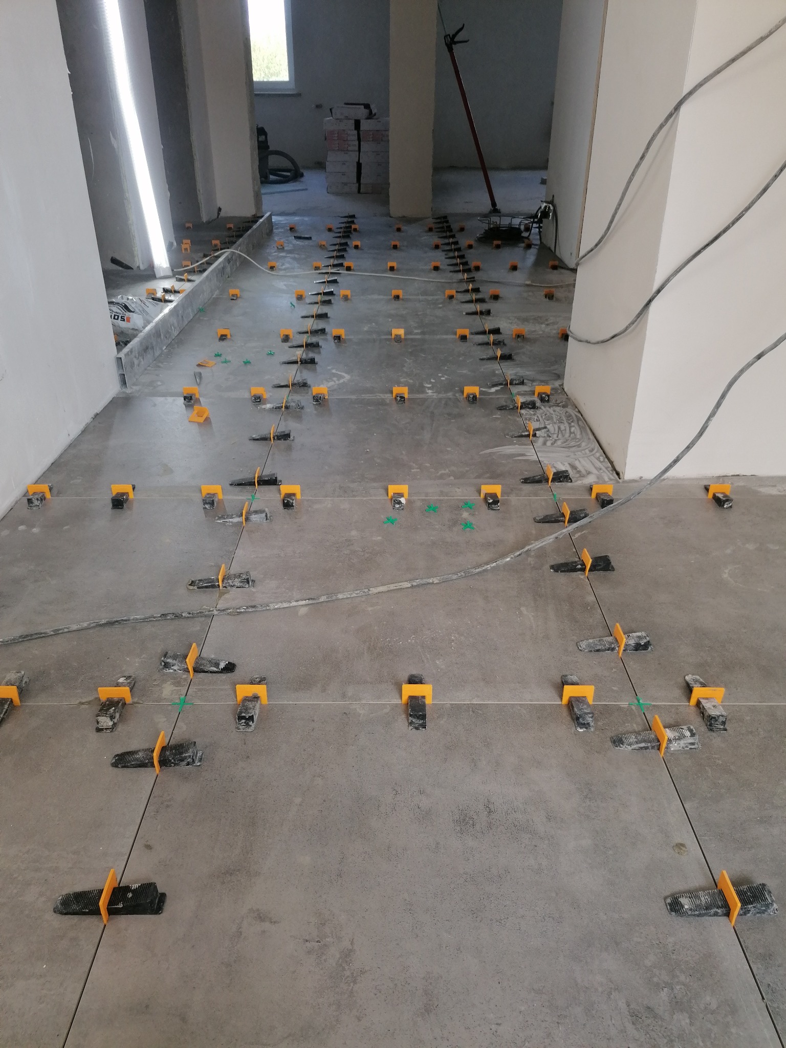 Szare płytki podłogowe ułożone z użyciem systemu poziomowania płytek z żółtymi klinami i czarnymi klipsami, widoczne zielone krzyżyki dystansowe, w tle narzędzia i materiały budowlane.