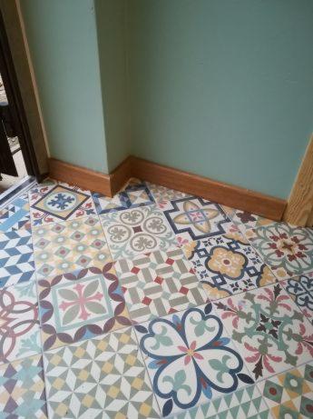 Wzorzysta, kolorowa podłoga wyłożona płytkami ceramicznymi w stylu patchwork, z drewnianymi listwami przypodłogowymi przy ścianie pomalowanej na kolor morski.