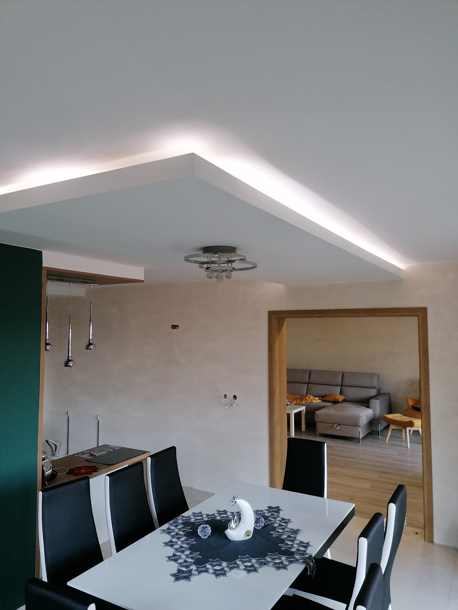 Nowoczesne wnętrze jadalni z podwieszanym sufitem i oświetleniem LED, widok na salon przez drewniane futryny, biały stół z czarnymi krzesłami i dekoracją w kształcie księżyca.