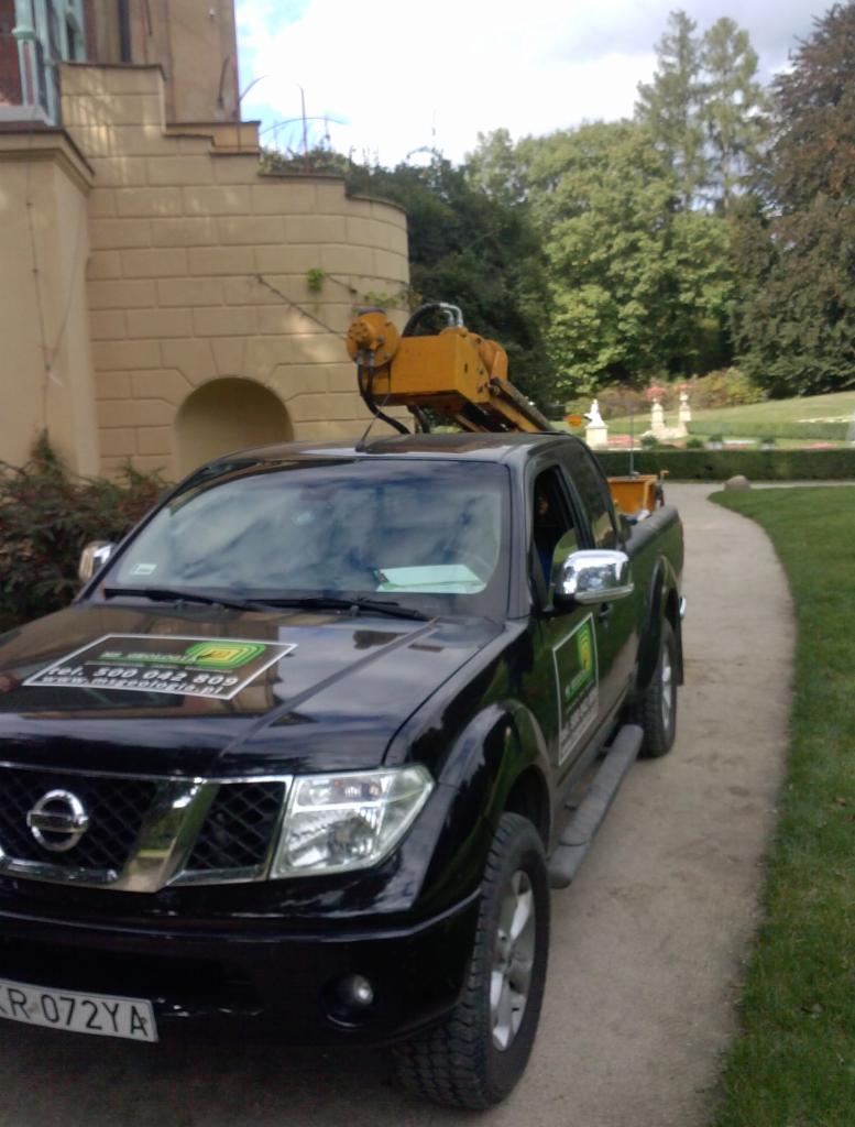 Czarny pickup Nissan z żółtym sprzętem do wierceń na pace, zaparkowany obok ścieżki w parku, z budynkiem w tle.