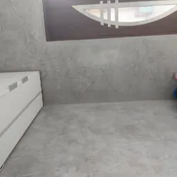 Szara, dekoracyjna ściana z efektem betonu w pokoju. Widoczna biała komoda z szufladami i ozdobny element nad oknem. Minimalistyczny wystrój wnętrza.