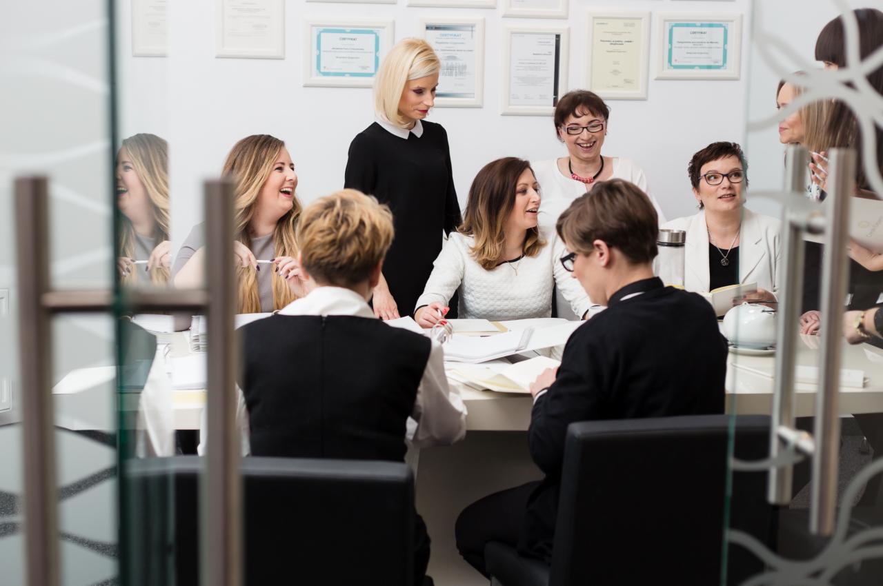 Zespół osób w biurze, siedzących przy białym stole podczas spotkania, w tle ściana z oprawionymi certyfikatami.