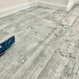 Szare panele podłogowe imitujące drewno, z widocznym opakowaniem paneli Classen Megaloc w rogu pomieszczenia, przy białej listwie przypodłogowej.