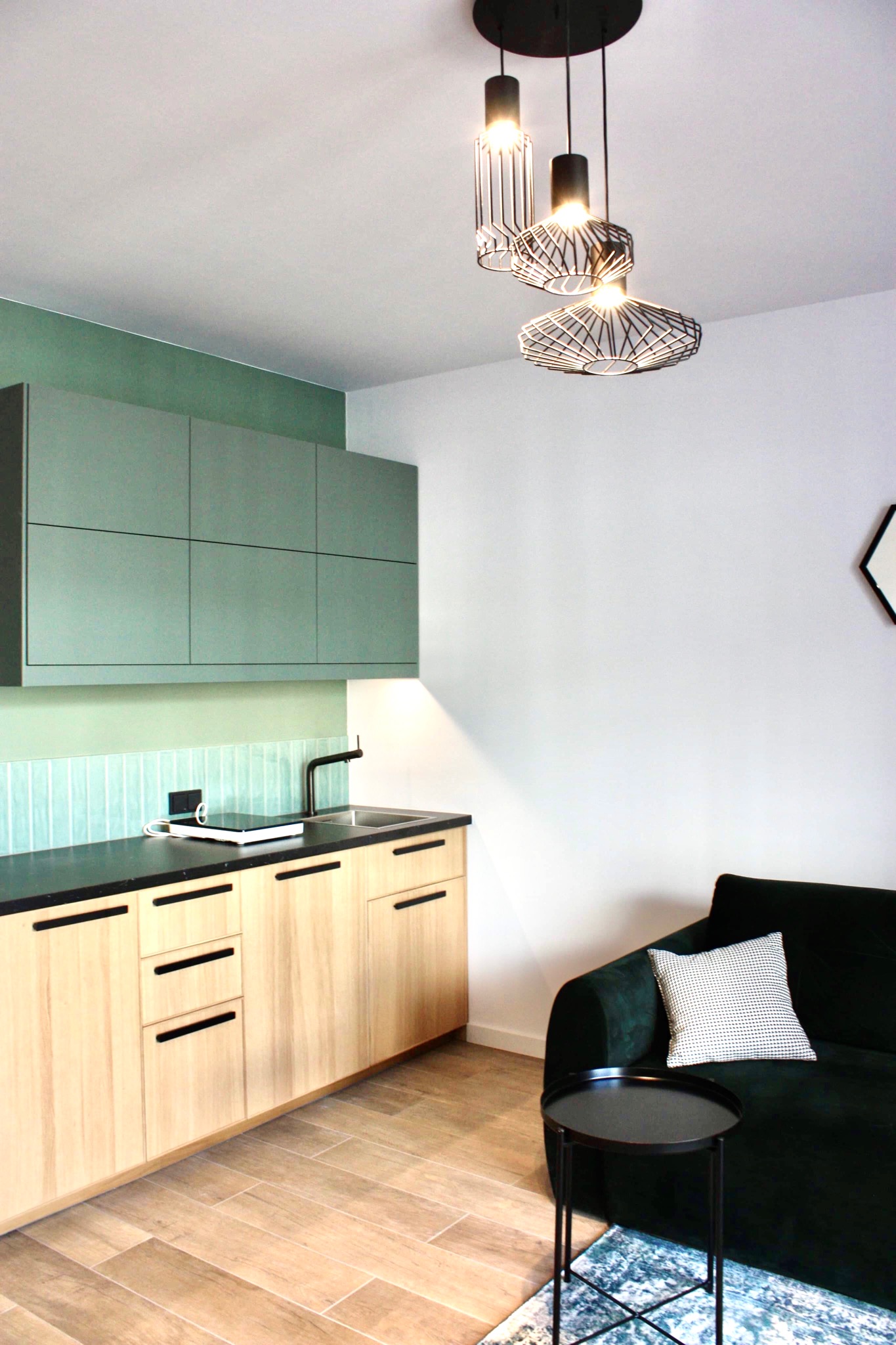 Nowoczesna kuchnia z drewnianymi szafkami, czarnym blatem i zielonymi akcentami, oświetlona designerskimi lampami wiszącymi, z widocznym fragmentem zielonej sofy i stolika kawowego.