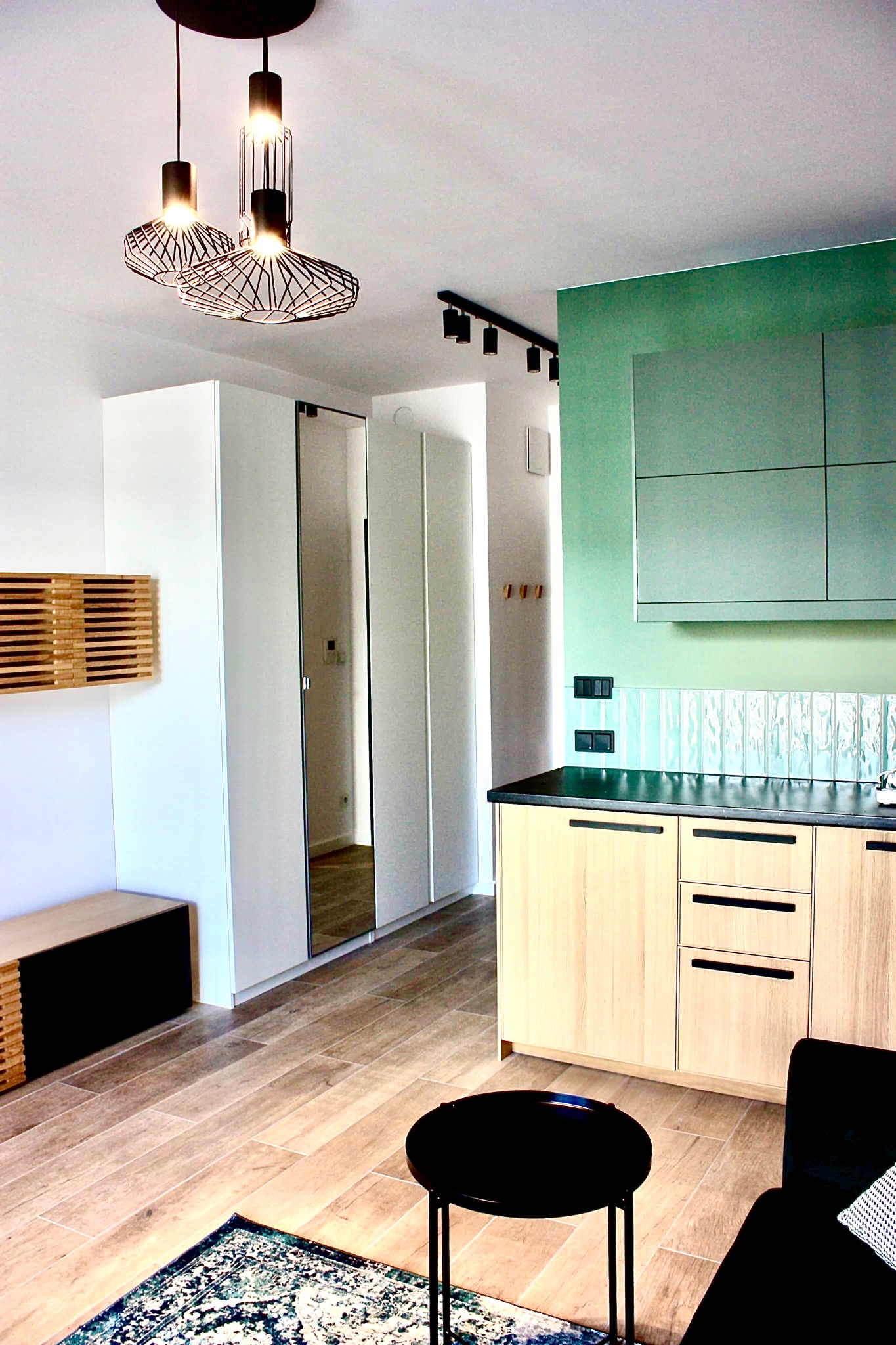 Nowoczesne wnętrze mieszkania z drewnianą podłogą, zieloną ścianą w kuchni, geometrycznymi lampami wiszącymi i czarnym stolikiem kawowym na wzorzystym dywanie.