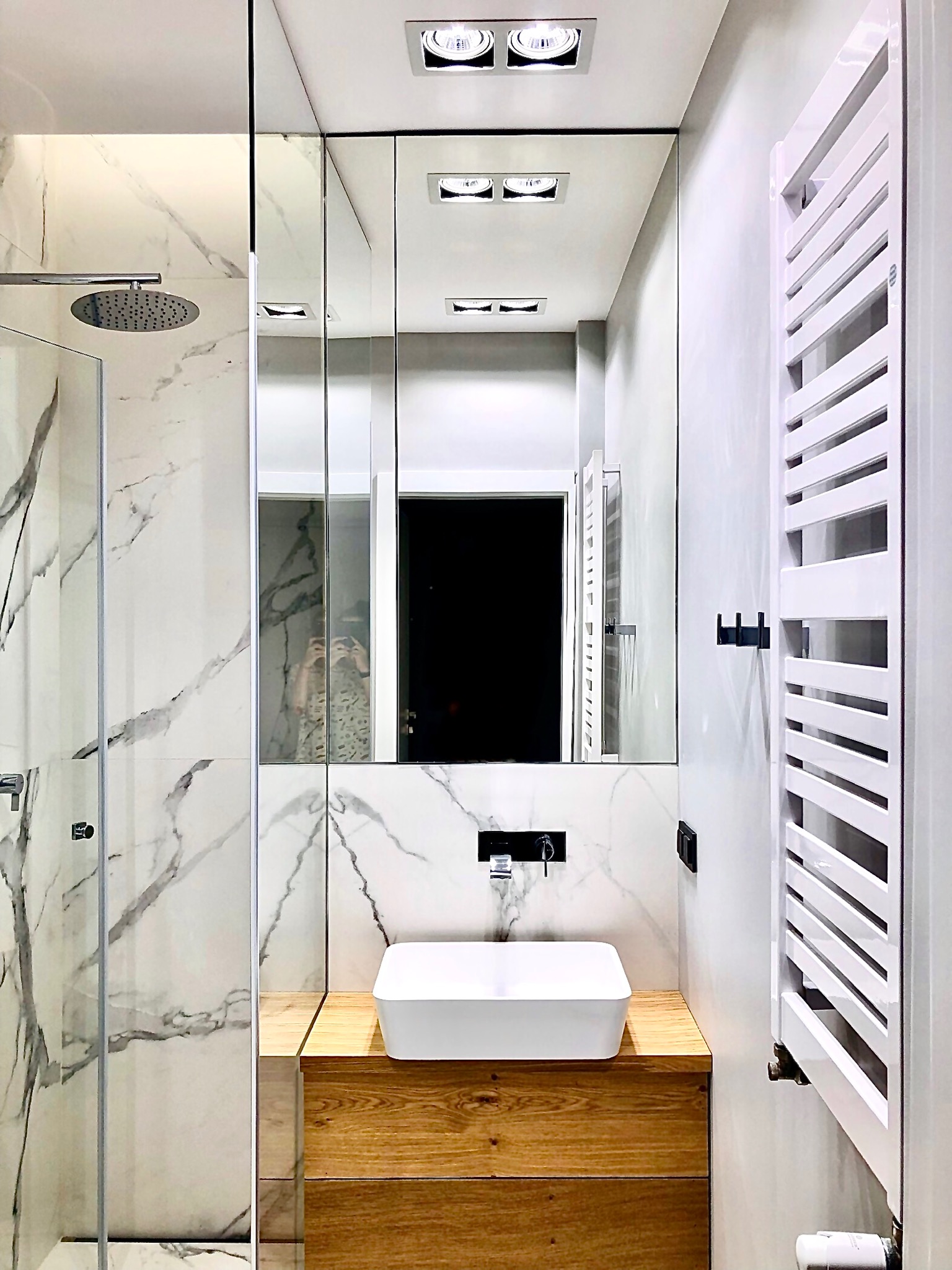 Nowoczesna łazienka z marmurowymi płytkami, drewnianą szafką pod umywalką nablatową, grzejnikiem drabinkowym i kabiną prysznicową typu walk-in. W lustrze odbija się osoba robiąca zdjęcie.