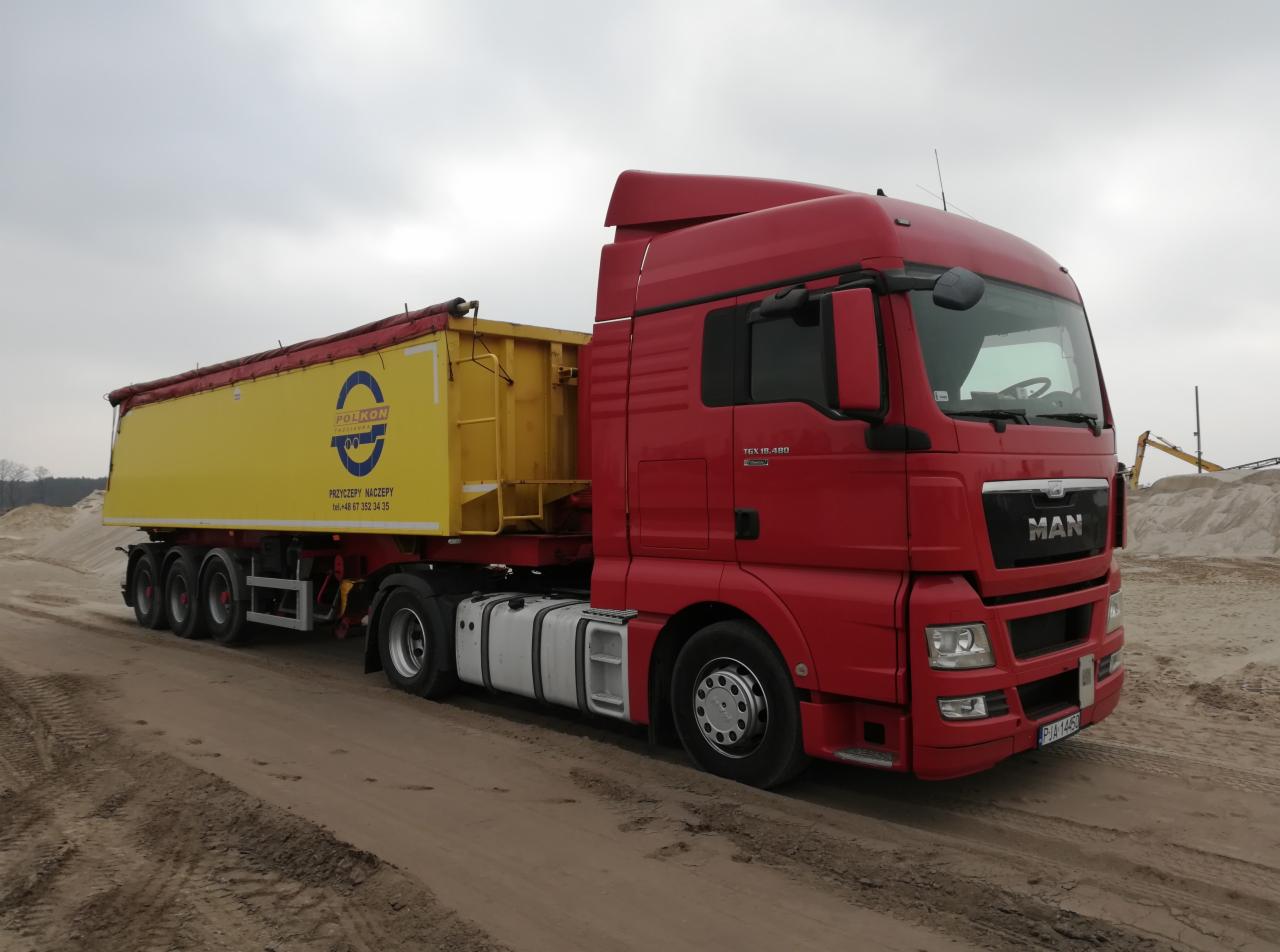 Czerwony ciągnik MAN z żółtą naczepą z logo Polkon stoi na piaszczystym terenie, w tle widoczna koparka.