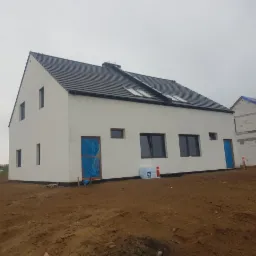 Surowy, biały dom jednorodzinny z czarną dachówką, widoczne zakryte folią drzwi wejściowe i okna, obok budynek w stanie surowym, ziemia wokół.