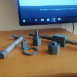 Szare, plastikowe komponenty mechaniczne, prawdopodobnie prototypy lub modele, wydrukowane w 3D, leżące na drewnianym biurku przed włączonym monitorem.