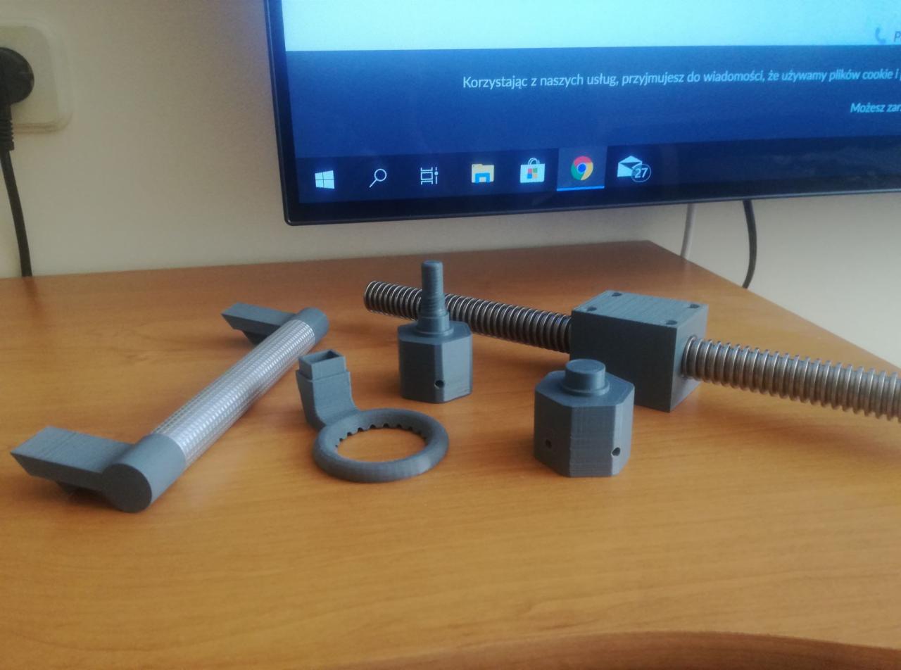 Szare, plastikowe komponenty mechaniczne, prawdopodobnie prototypy lub modele, wydrukowane w 3D, leżące na drewnianym biurku przed włączonym monitorem.